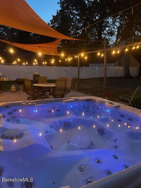 pool at night with a hot tub, a patio, and fence