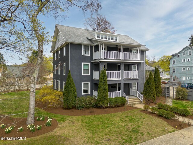 view of front of house with roof with shingles, a front lawn, a chimney, a balcony, and covered porch
