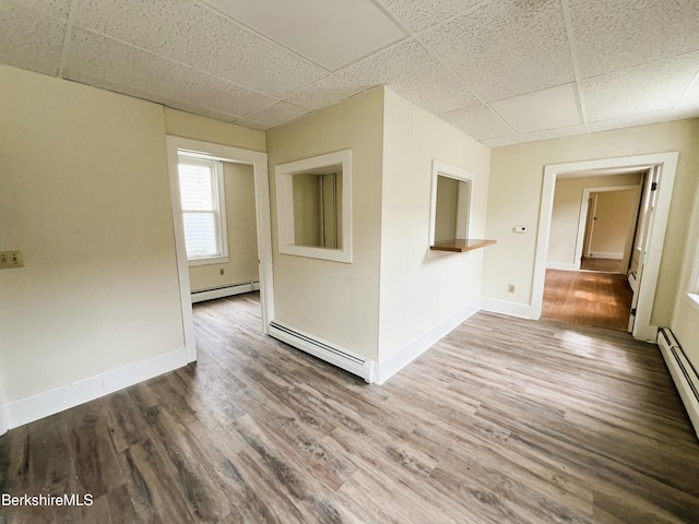 empty room with a baseboard heating unit, wood finished floors, a drop ceiling, and baseboards