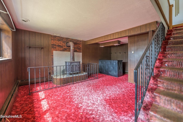 carpeted living room featuring stairway, wood walls, a wood stove, and a baseboard radiator