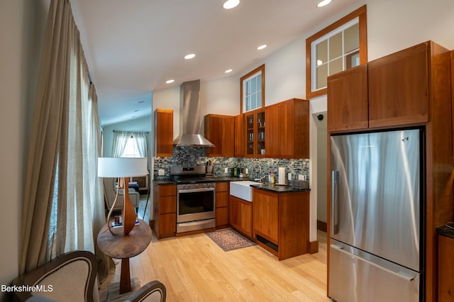 kitchen featuring tasteful backsplash, brown cabinetry, dark countertops, wall chimney exhaust hood, and appliances with stainless steel finishes