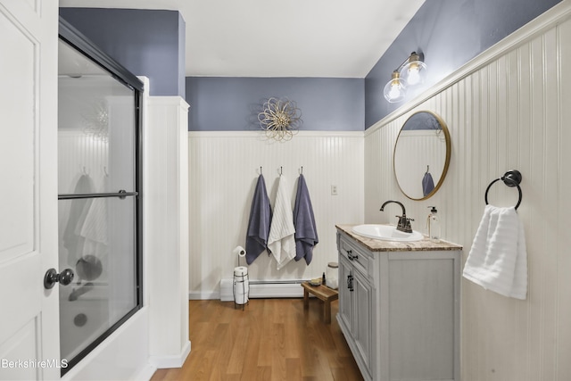 bathroom featuring wood-type flooring, vanity, wooden walls, bath / shower combo with glass door, and a baseboard heating unit