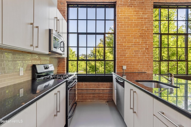 kitchen featuring sink, dark stone countertops, appliances with stainless steel finishes, white cabinetry, and brick wall