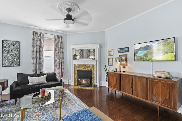living area with ornamental molding, a glass covered fireplace, baseboards, ceiling fan, and dark wood-type flooring