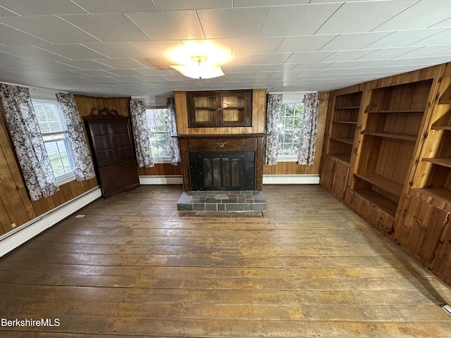 unfurnished living room with a baseboard heating unit, hardwood / wood-style flooring, a glass covered fireplace, built in features, and wooden walls
