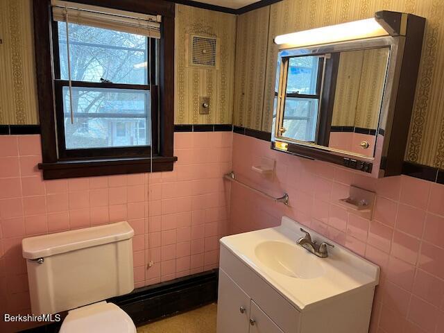 half bath featuring wallpapered walls, toilet, vanity, and a wainscoted wall