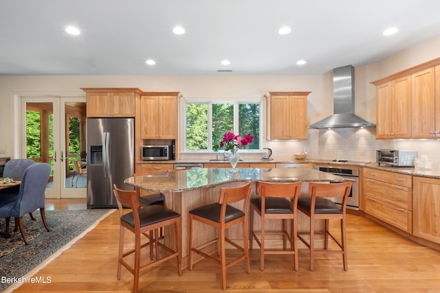 kitchen with a center island, wall chimney exhaust hood, a kitchen breakfast bar, light stone counters, and appliances with stainless steel finishes