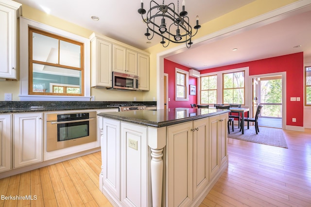 kitchen with appliances with stainless steel finishes, an inviting chandelier, light wood finished floors, dark stone counters, and a kitchen island
