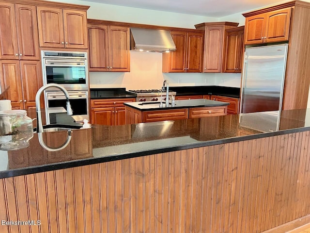 121 E Dugway Rd, Lenox, MA 01240 - kitchen featuring wall chimney range hood, appliances with stainless steel finishes, an island with sink, brown cabinetry, and dark stone countertops