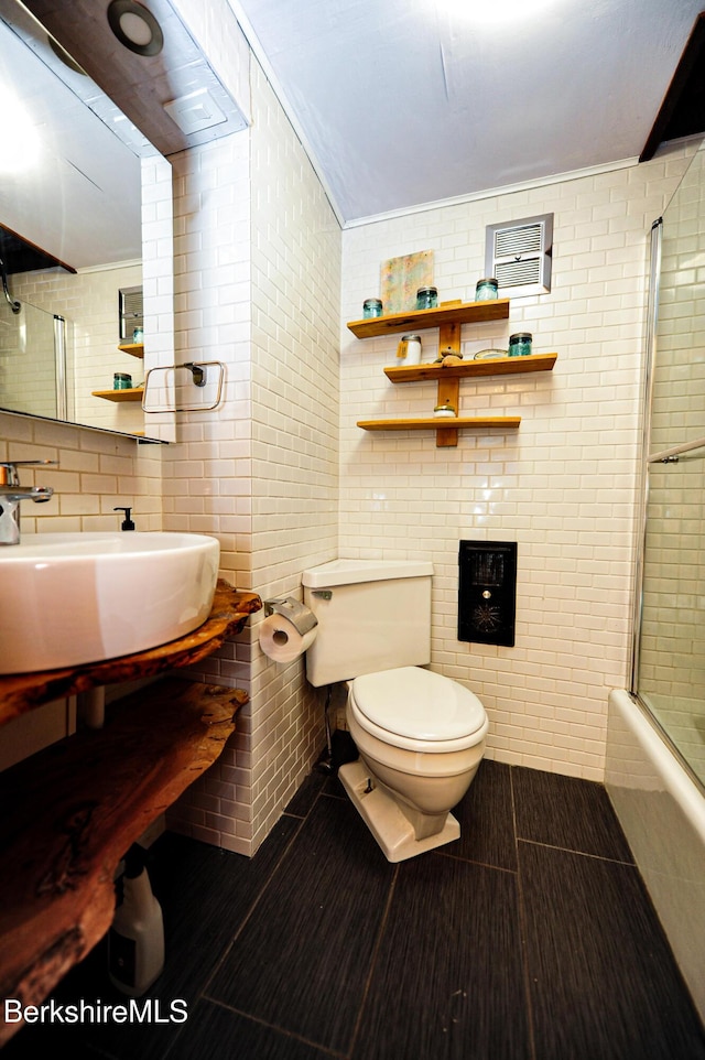 bathroom featuring wood finished floors, shower / bath combination with glass door, a sink, tile walls, and toilet