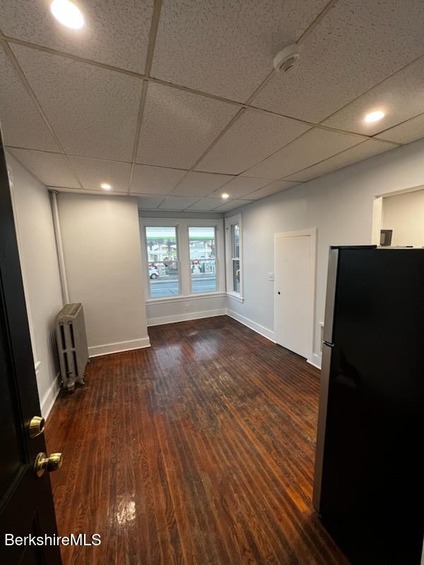 empty room with baseboards, a drop ceiling, dark wood finished floors, and radiator