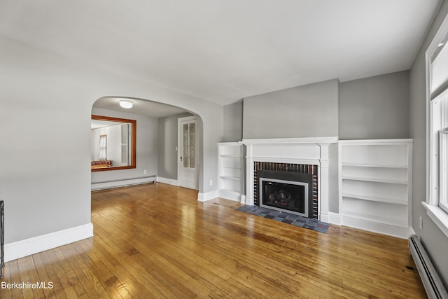 unfurnished living room with a brick fireplace, a baseboard heating unit, hardwood / wood-style floors, arched walkways, and a baseboard radiator