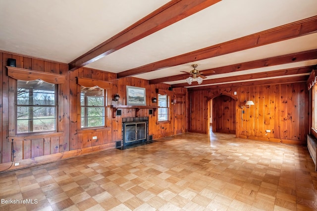 unfurnished living room with wood walls, arched walkways, beam ceiling, a fireplace with flush hearth, and baseboards