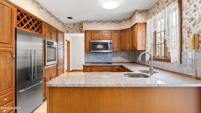 kitchen featuring a sink, a peninsula, brown cabinets, light stone countertops, and appliances with stainless steel finishes