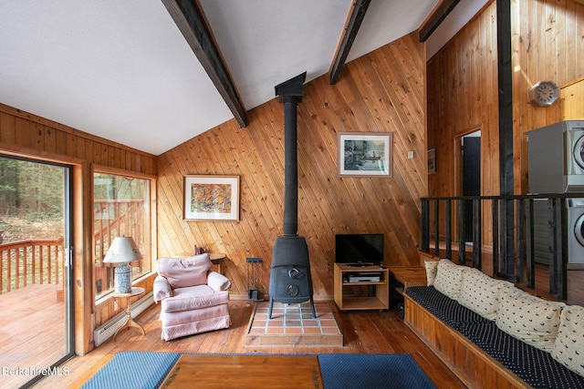 living area with a wood stove, a baseboard radiator, stacked washer and clothes dryer, lofted ceiling with beams, and wooden walls