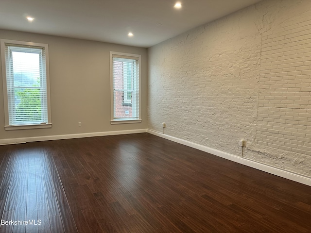 spare room with healthy amount of natural light, baseboards, dark wood finished floors, recessed lighting, and brick wall