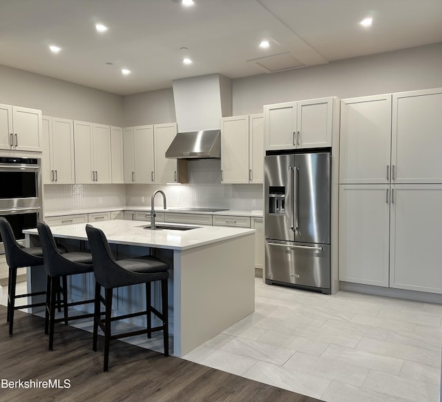 kitchen with stainless steel appliances, wall chimney range hood, a sink, a center island with sink, and decorative backsplash