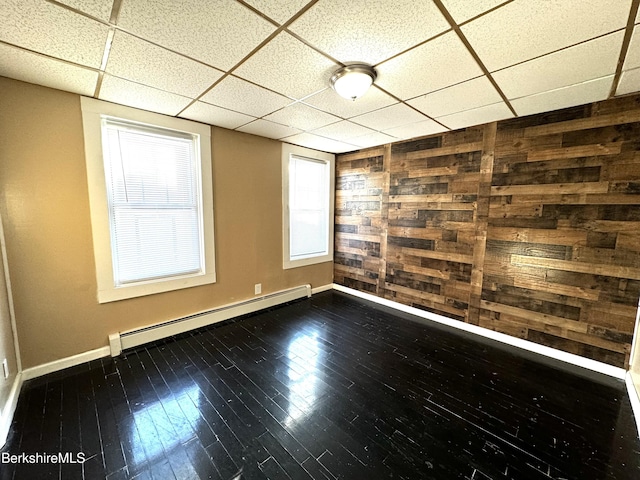 empty room featuring hardwood / wood-style flooring, a paneled ceiling, a healthy amount of sunlight, a baseboard radiator, and wood walls