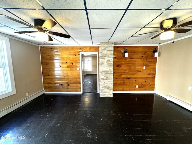 empty room with ceiling fan, a baseboard heating unit, wooden walls, and a drop ceiling