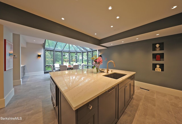 kitchen featuring sink, built in shelves, an island with sink, dark brown cabinets, and light stone counters