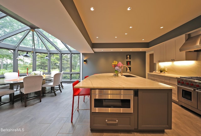 kitchen with gray cabinetry, sink, wall chimney exhaust hood, luxury stove, and a kitchen island with sink