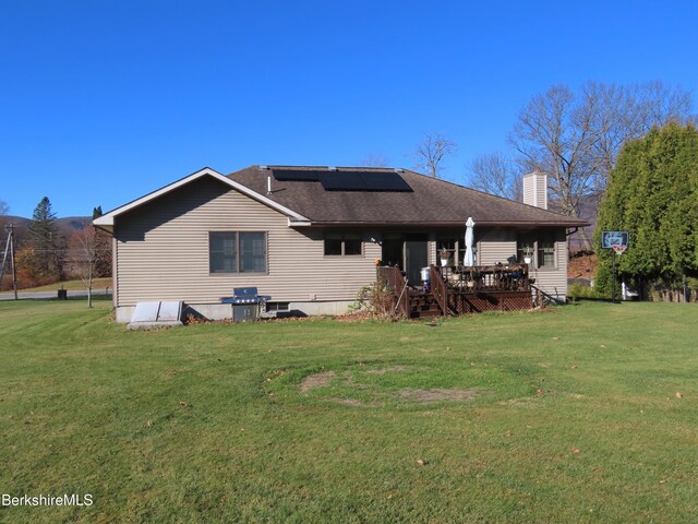 rear view of property with solar panels, a wooden deck, roof with shingles, a chimney, and a yard