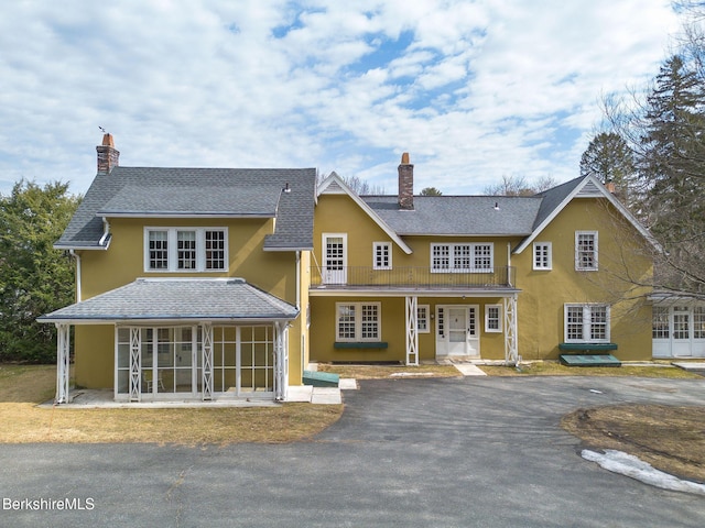 shingle-style home featuring a shingled roof, stucco siding, a balcony, and a chimney