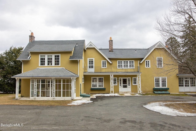 view of front of house with a balcony, a sunroom, a shingled roof, stucco siding, and a chimney