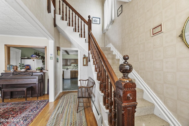 staircase with wood finished floors, baseboards, wallpapered walls, a towering ceiling, and crown molding