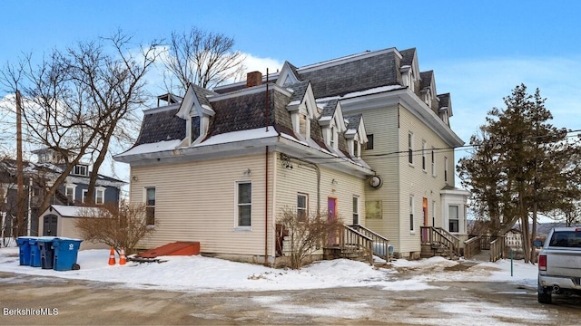 view of snowy exterior featuring a shingled roof, mansard roof, and a chimney