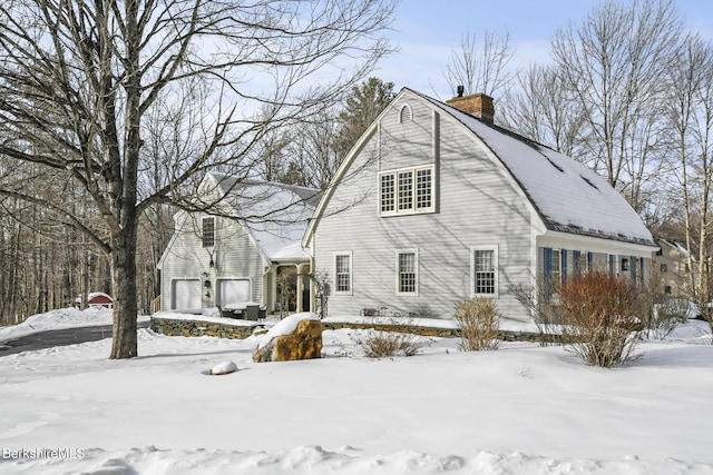 snow covered property featuring a garage, a chimney, and a gambrel roof