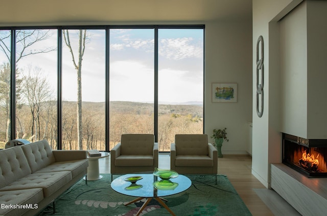 living room with baseboards, a forest view, wood finished floors, a wall of windows, and a multi sided fireplace