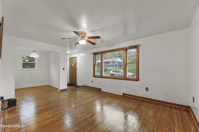 empty room featuring arched walkways, ceiling fan, baseboard heating, hardwood / wood-style flooring, and baseboards