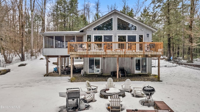 snow covered rear of property featuring a wooden deck, a sunroom, and an outdoor fire pit