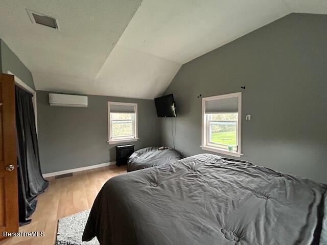 bedroom with a wall mounted air conditioner, lofted ceiling, light wood-type flooring, and multiple windows