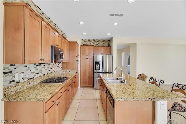 kitchen featuring an island with sink, stainless steel appliances, light tile floors, a kitchen bar, and light stone countertops