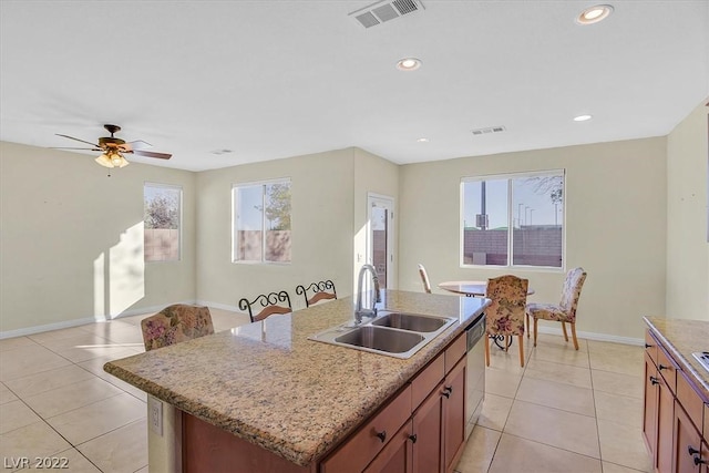 kitchen with ceiling fan, sink, light tile floors, light stone counters, and a center island with sink