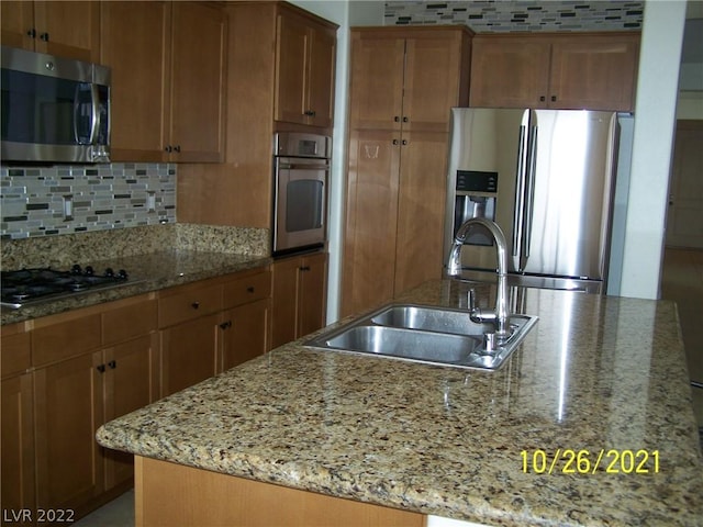 kitchen featuring stainless steel appliances, light stone countertops, a center island with sink, backsplash, and sink