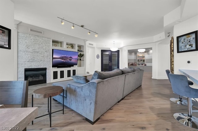 living room featuring built in features, a stone fireplace, rail lighting, and light hardwood / wood-style flooring