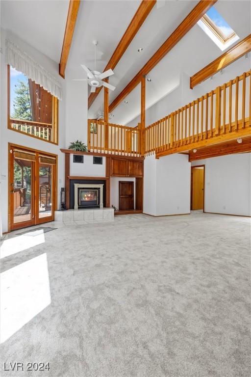 unfurnished living room with a tiled fireplace, beam ceiling, light colored carpet, ceiling fan, and a towering ceiling