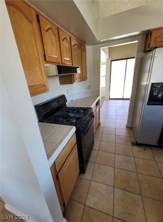 kitchen with tile counters, stainless steel fridge, black stove, range hood, and light tile flooring