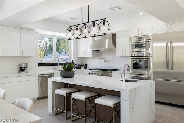 kitchen featuring wall chimney exhaust hood, an island with sink, decorative light fixtures, and appliances with stainless steel finishes