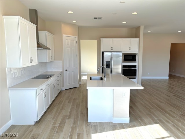 kitchen with appliances with stainless steel finishes, wall chimney range hood, an island with sink, white cabinetry, and light hardwood / wood-style floors