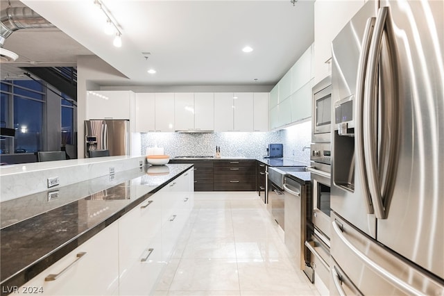 kitchen with light tile flooring, dark stone counters, appliances with stainless steel finishes, white cabinets, and backsplash