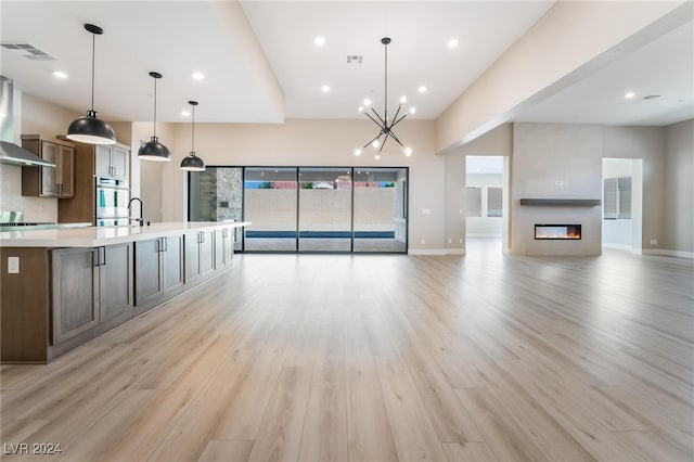 kitchen with a large island with sink, light wood-type flooring, double oven, a large fireplace, and decorative light fixtures