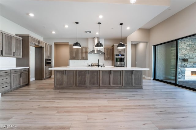 kitchen with wall chimney exhaust hood, pendant lighting, light hardwood / wood-style flooring, stainless steel oven, and a large island