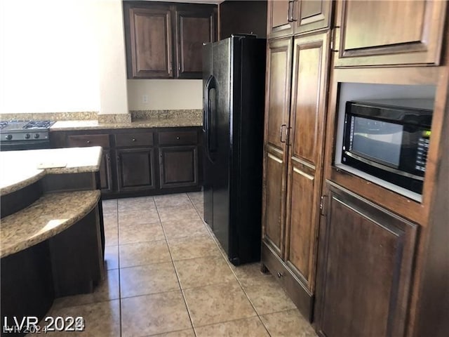 kitchen with dark brown cabinetry, light stone counters, light tile flooring, and black appliances