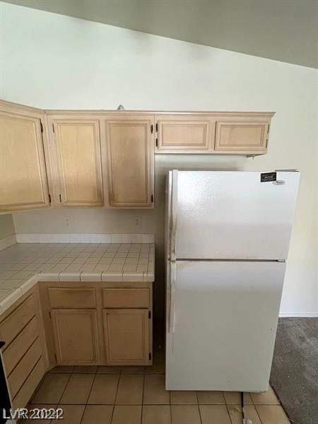 kitchen with light tile flooring, white refrigerator, light brown cabinets, vaulted ceiling, and tile countertops