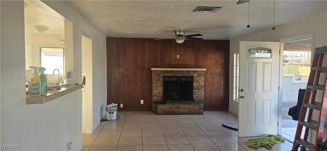 living room with ceiling fan, a stone fireplace, light tile patterned flooring, wooden walls, and visible vents