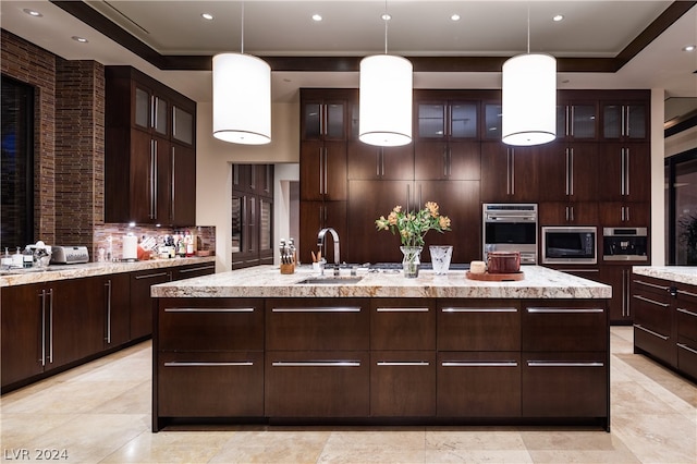 kitchen featuring decorative light fixtures, backsplash, light tile flooring, an island with sink, and dark brown cabinetry